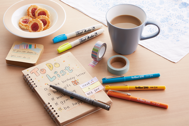 Artline markers and pens beside a hand-lettered notebook